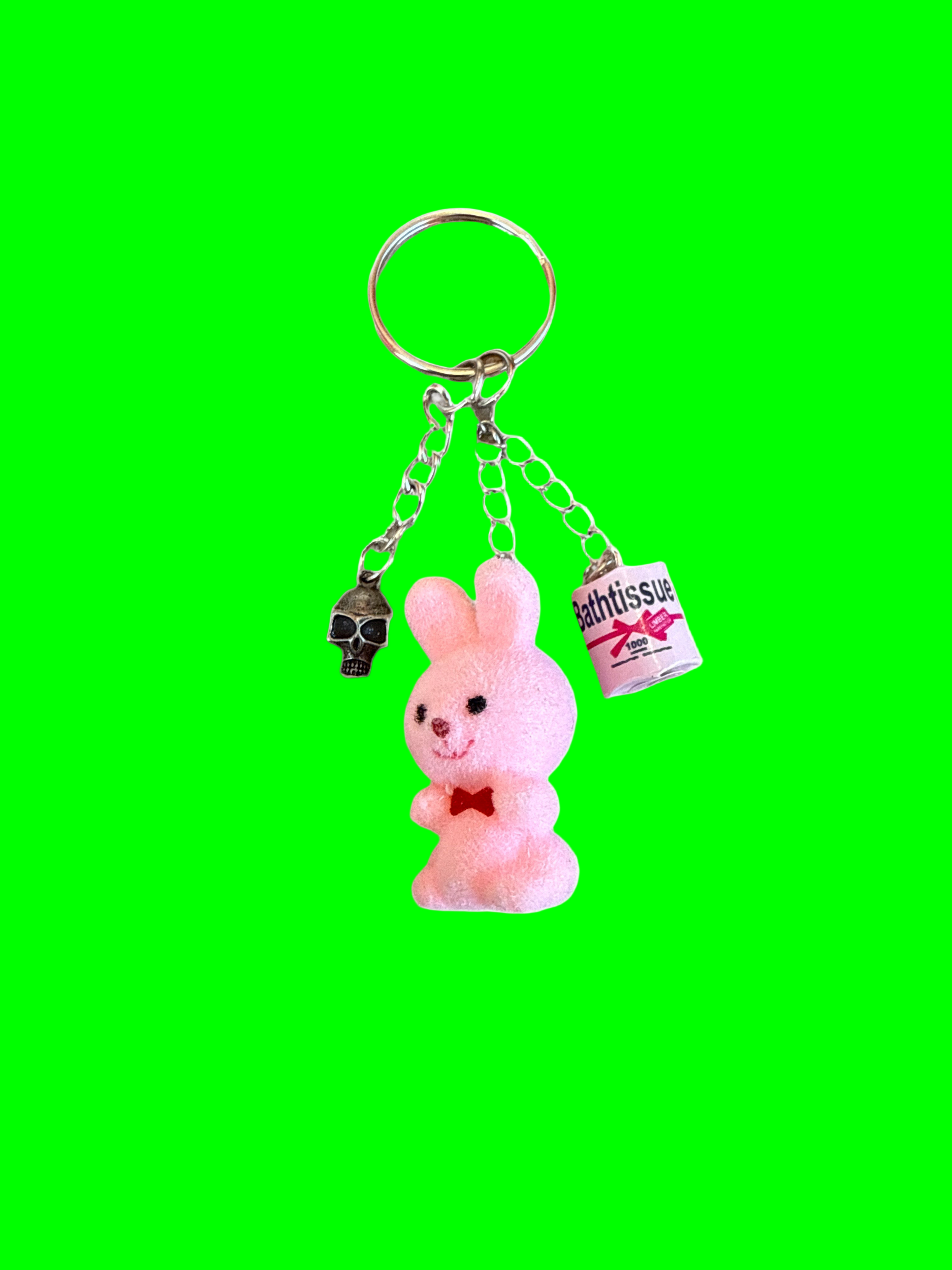 Keychain with pink rabbit charm, skull, and toilet paper roll on a green background
